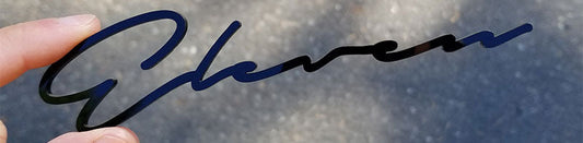 Gloss black Eleven script automotive emblem in flowing signature-style lettering, precision-cut acrylic construction held against outdoor background.