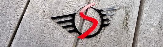 Custom Mini Cooper winged S emblem in black with red center made from UV rated acrylic displayed on wooden surface.