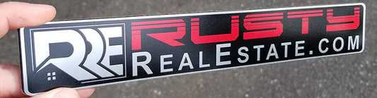 Custom real estate automotive badge with white and red lettering on black background, precision-cut layered acrylic construction, held horizontally for size reference.