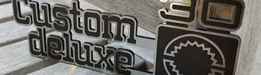 Custom Deluxe 30 automotive emblem with brushed metallic raised lettering, black layered backing, and square accent badge, precision-cut stacked construction displayed on wood surface.