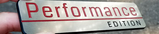 Performance Edition automotive emblem with brushed silver background, red Performance lettering, black Edition text, red underline accent, and layered black backing held in hand.
