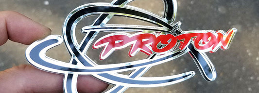 Chrome Proton emblem with red script and black accents surrounded by chrome orbital design made from UV rated acrylic held outdoors.