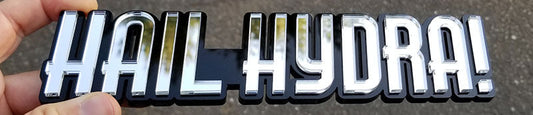Chrome-style Hail Hydra automotive emblem with black backing made from UV-rated acrylic held in hand outdoors.