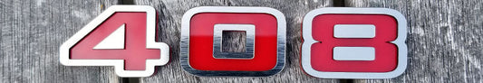 Brushed stainless steel and red 408 automotive emblem with layered numeric design displayed on wooden surface.