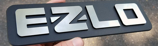EZ-LO automotive emblem with brushed silver raised lettering on matte black rectangular backing, precision-cut layered acrylic construction held in hand.
