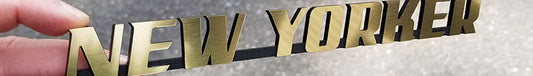 Brushed gold New Yorker automotive emblem with black backing made from UV-rated acrylic held in hand outdoors.