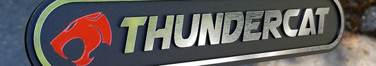 Thundercat automotive badge with brushed metallic lettering, red cat head logo, olive accent border, and layered black panel held in hand.