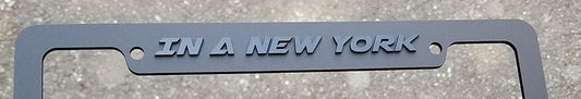 Matte black wide #3 license plate frame with gloss black Racer font lettering reading In a New York State of Mind held outdoors.