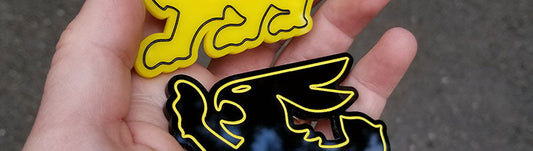 Two stacked Mean Bunny automotive emblems in gloss black with yellow outline and solid yellow with black detailing, layered acrylic construction, handheld showing reflective finish and smooth edges.