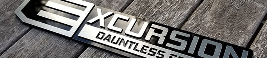 Excursion Dauntless Edition automotive emblem with brushed silver lettering, black layered backing, and stacked acrylic construction displayed on weathered wood surface.