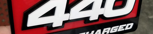 440 Supercharged automotive emblem with large white numerals on red background, black border, and stacked acrylic layered construction held in hand.