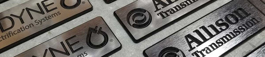 Allison and Odyne brushed stainless acrylic corporate badges with beveled edges and layered business logos arranged on metal surface.