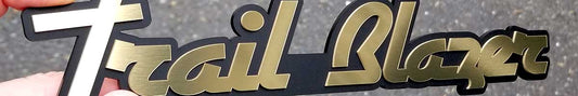 Gold brushed Trail Blazer automotive emblem with matte black backing, script lettering held in hand outdoors.
