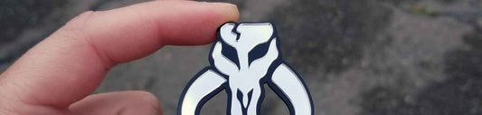 Small chrome horned skull automotive emblem with black outline and layered acrylic construction, reflective finish, held between fingers for size reference.