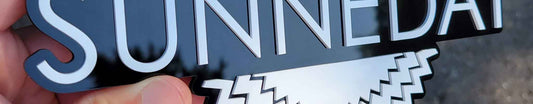 Gloss black and satin white Sunneday emblem with layered sunburst background and raised white lettering.