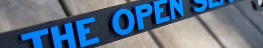 Satin black custom license plate frame with raised matte blue 3D lettering reading The Open Sea Is Where I’m Free.