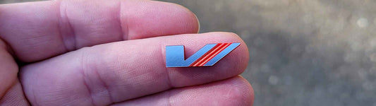 Small silver car emblem measuring 3/4 inch by 1/4 inch with diagonal red ridged engraved stripes, held on fingertips for size reference.