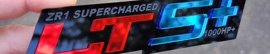 LTS+ ZR1 Supercharged performance badge with red chrome LT, blue chrome S+, and gloss black engraved backing made from UV rated acrylic held outdoors.
