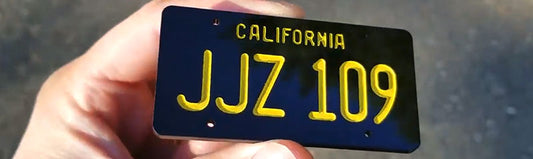 Miniature black license plate with engraved and painted yellow lettering reading JJZ 109 made from UV rated acrylic held outdoors.